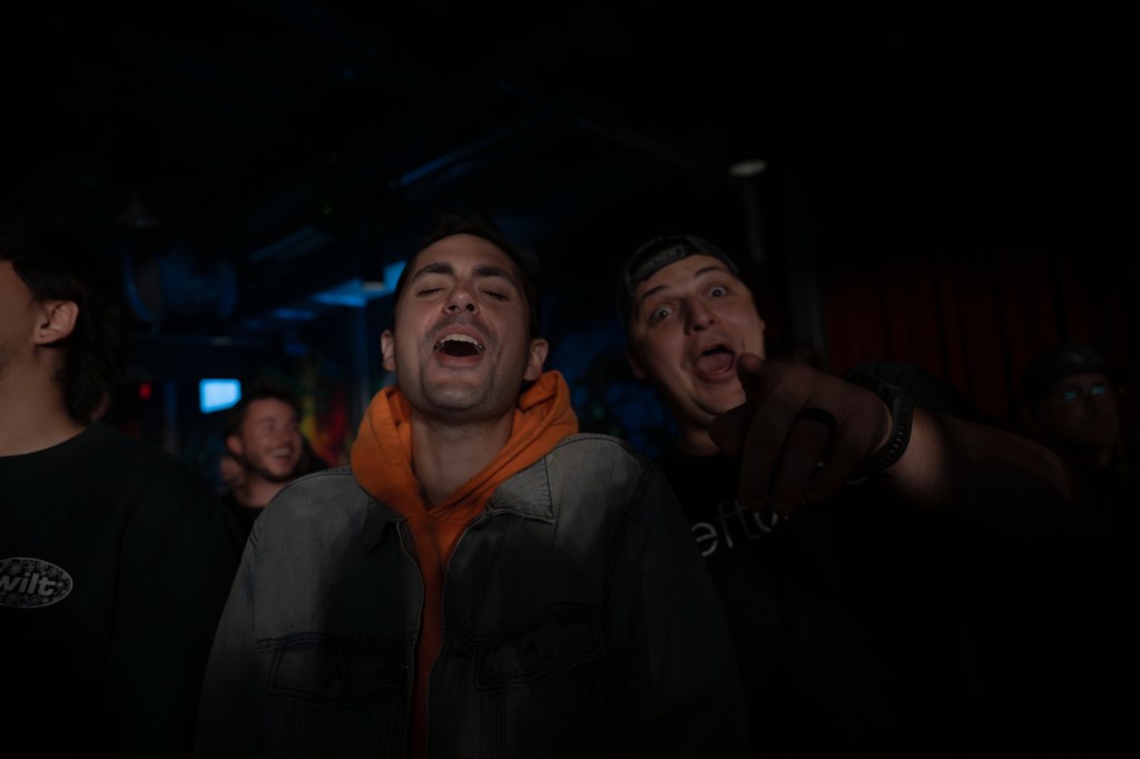 Two men singing passionately at a concert, with one wearing a denim jacket and an orange hoodie, while the other is enthusiastically pointing and smiling.